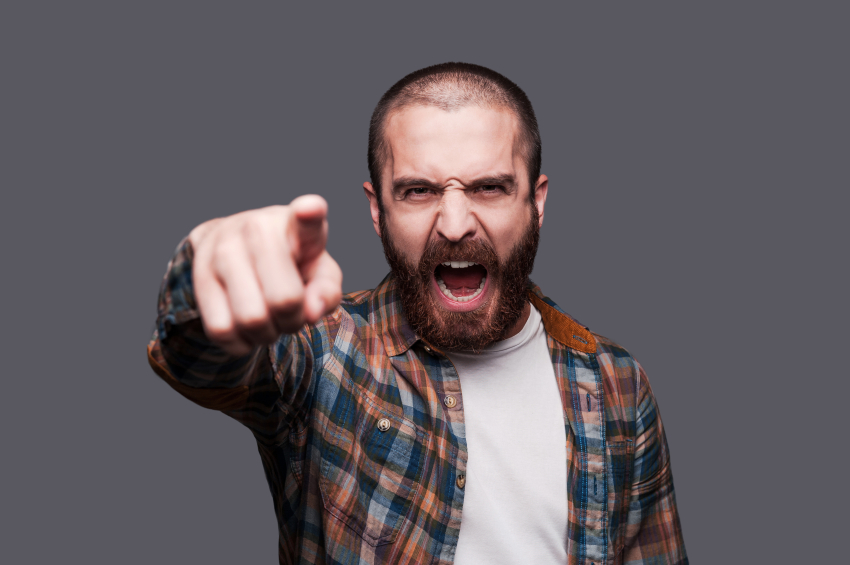Hey you! Furious young bearded man pointing you and shouting while standing against grey background