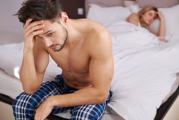Stressed man sits in bed whilst female sleeps