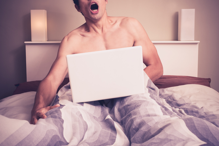 Young naked man is sitting in a bed and watching pornography on his laptop