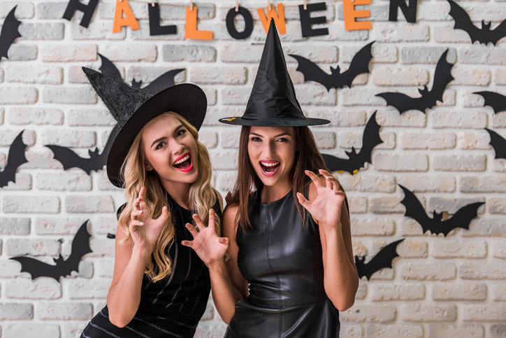 Beautiful girls in black dresses and witch hats are showing claws, looking at camera and smiling, on background decorated for Halloween