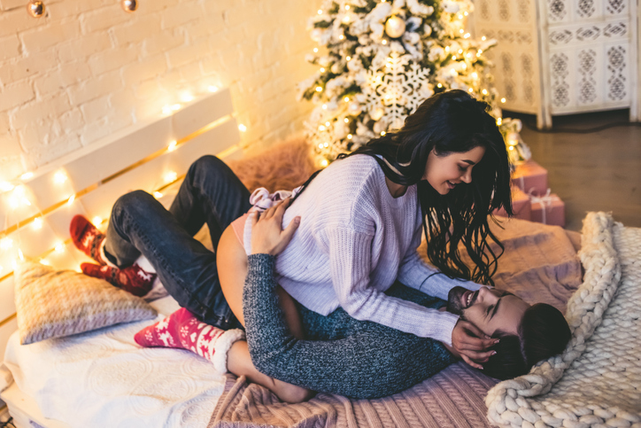 Passionate romantic couple spending time before New Year near beautiful Christmas tree at home. Sexy young woman and handsome man are lying on bed in sweaters and hugging. Tender couple sleep together