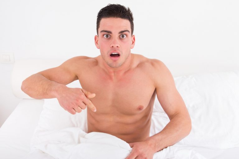 surprised and shocked half naked young man in bed looking down at his underwear at his penis under white covers sheet in badroom. Concept photo of male sexuality and man sex problems, domestic atmosphere.