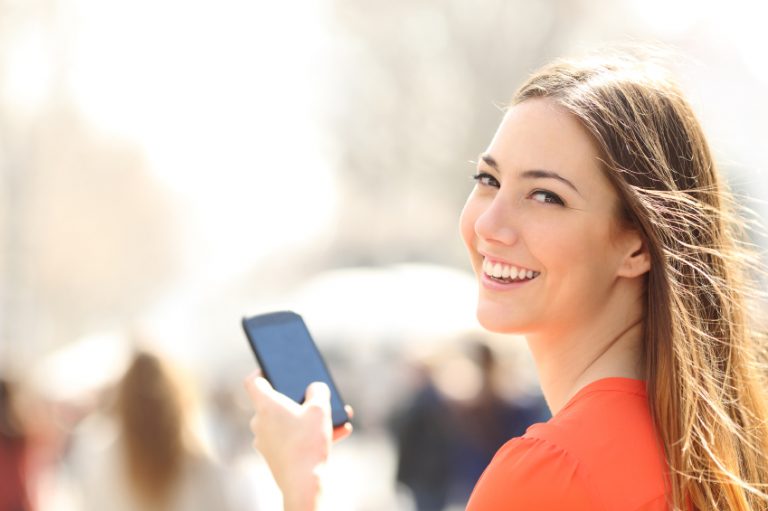Happy woman smiling and walking in the street using a smartphone and looking at camera
