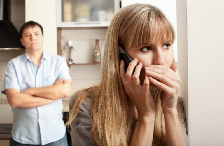Woman whispers on phone while man looks suspicious in the background